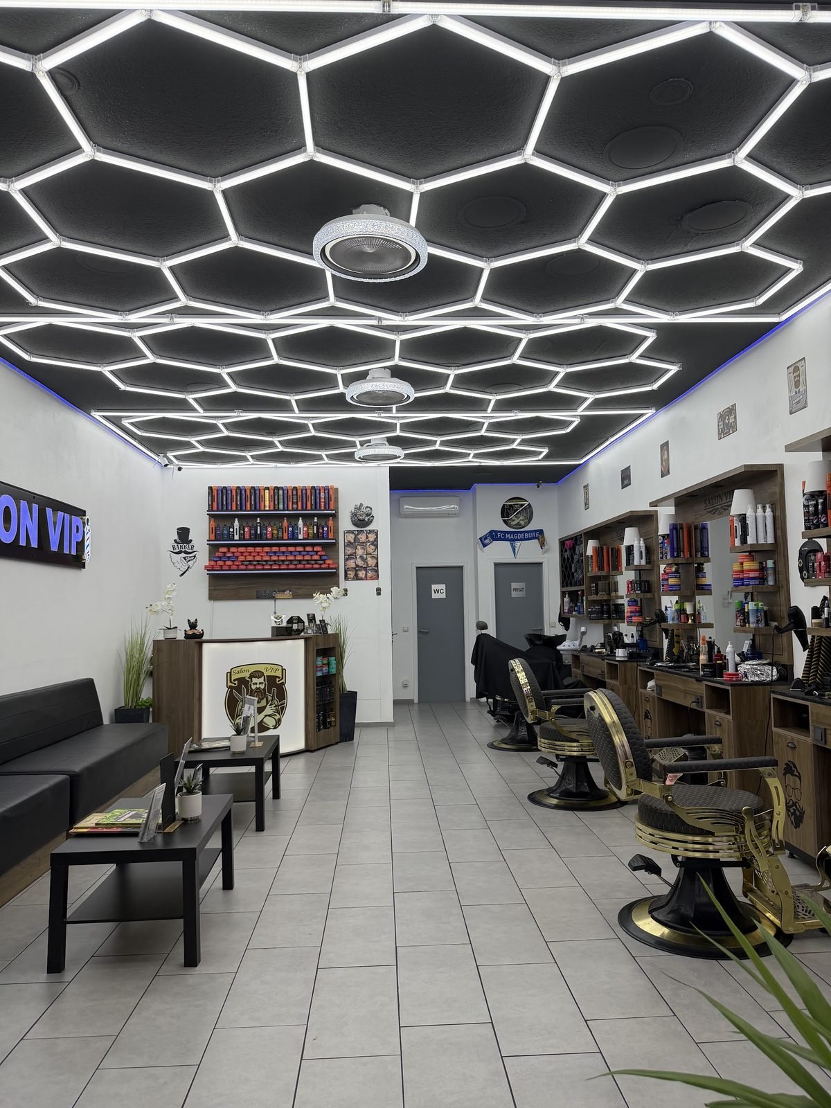 Blick der Länge nach durch den Salon VIP: Wabenlicht an der Decke, goldene Barberstühle rechts, Wartesofa links